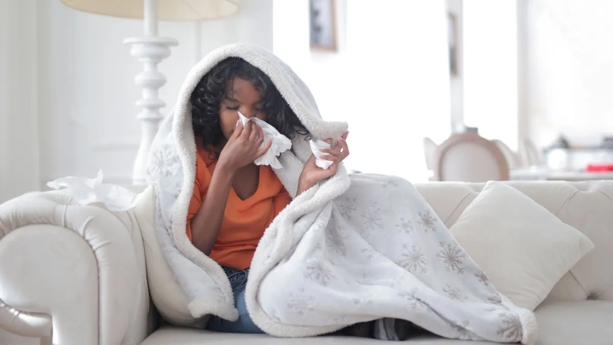 Mulher com gripe no sofá, coberta com manta e assoando o nariz