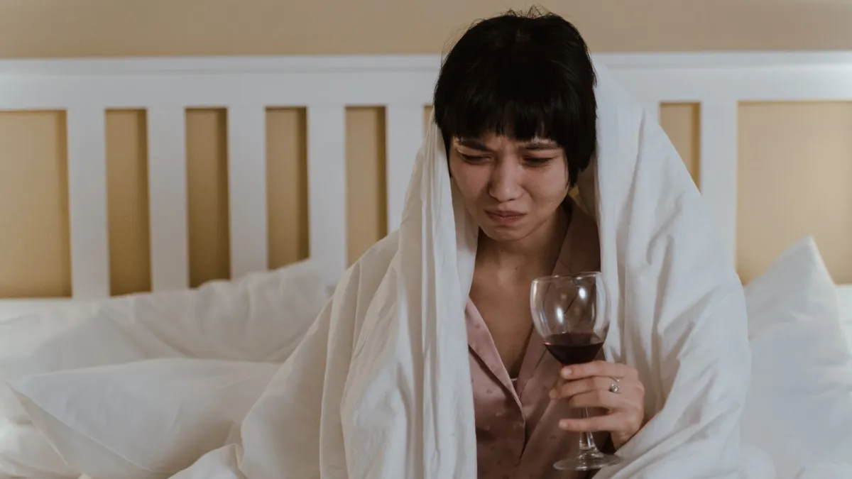Mulher de ressaca coberta com edredom segurando uma taça de vinho